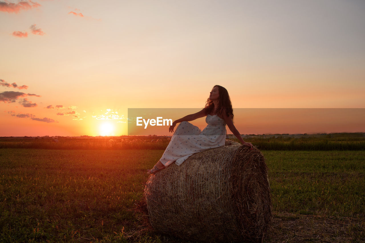 Woman sitting on haystack against sky during sunset