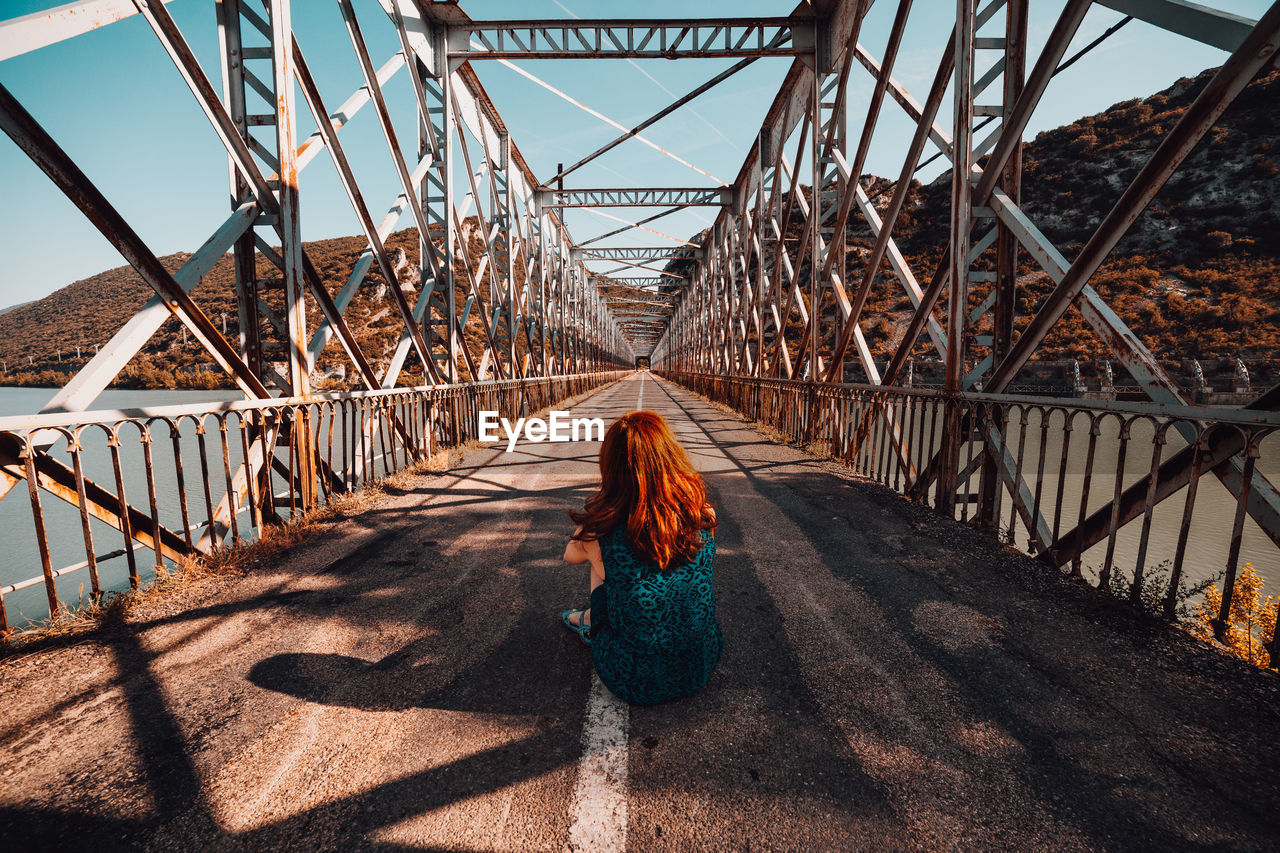 Rear view of woman sitting on footbridge against sky