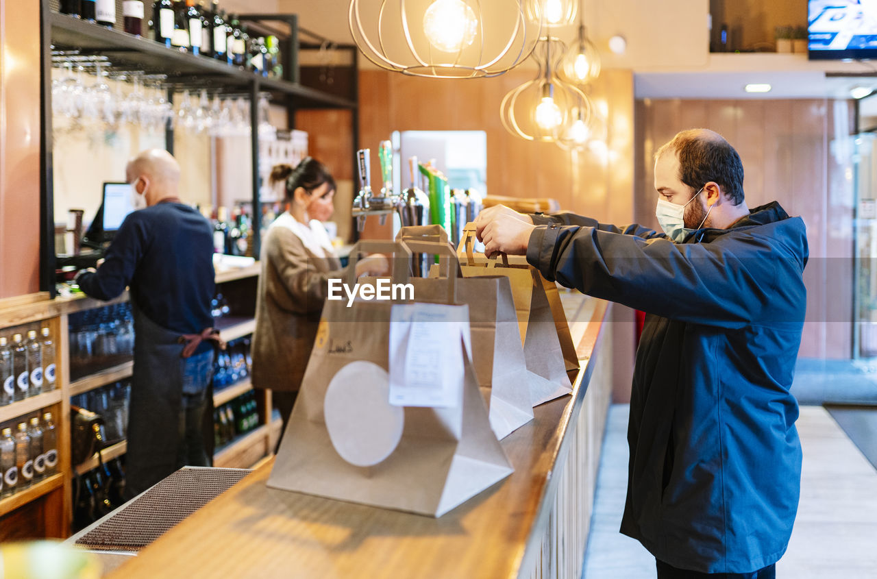 Young male customer picking up his order on bar counter while owners working in background during pandemic