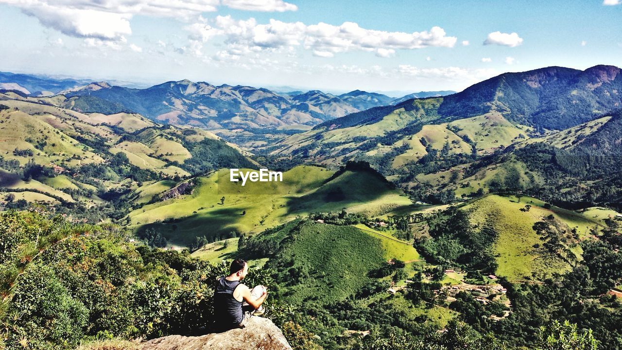 High angle view of male hiker sitting on cliff against mountains