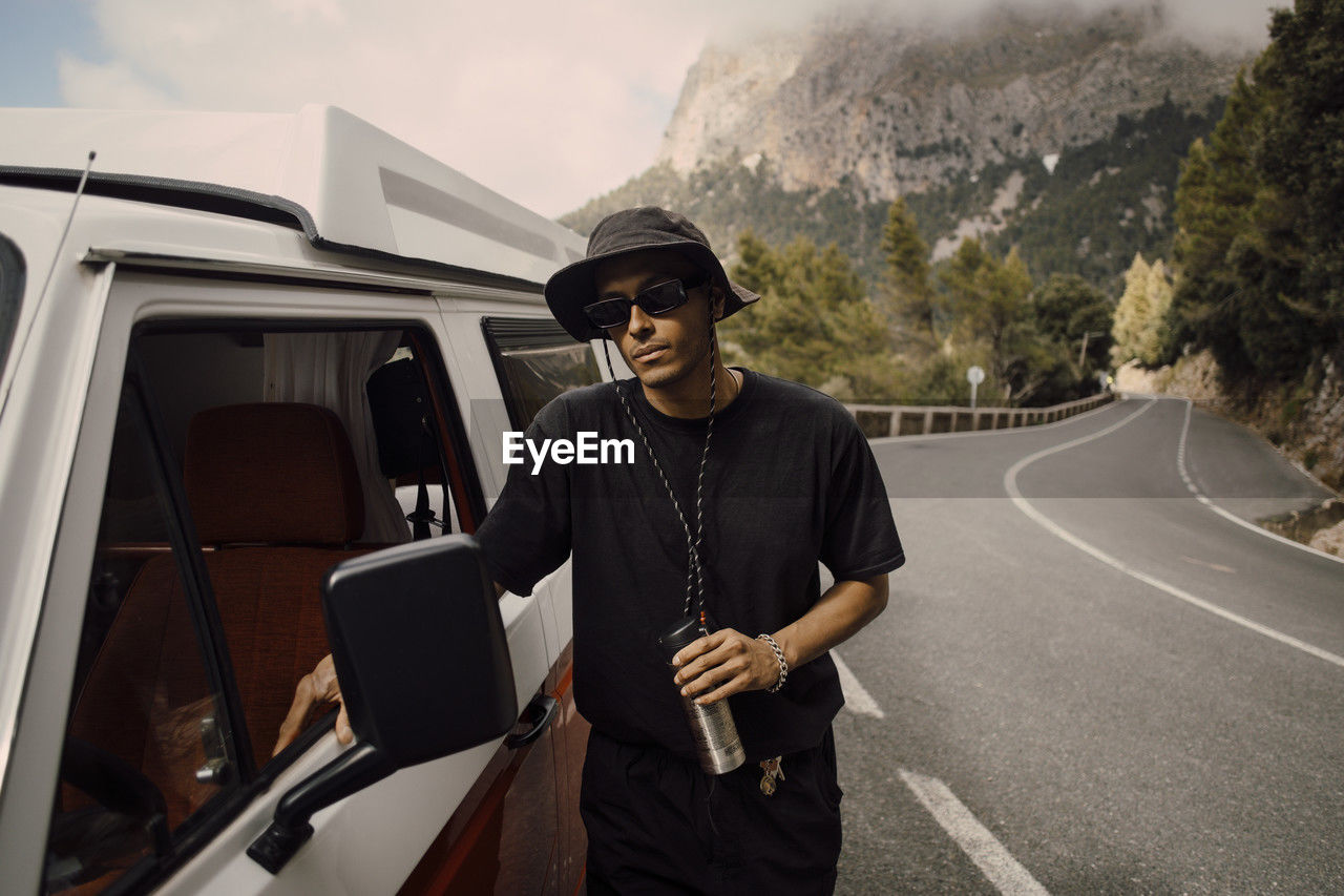 Young man wearing sunglasses standing next to van during road trip