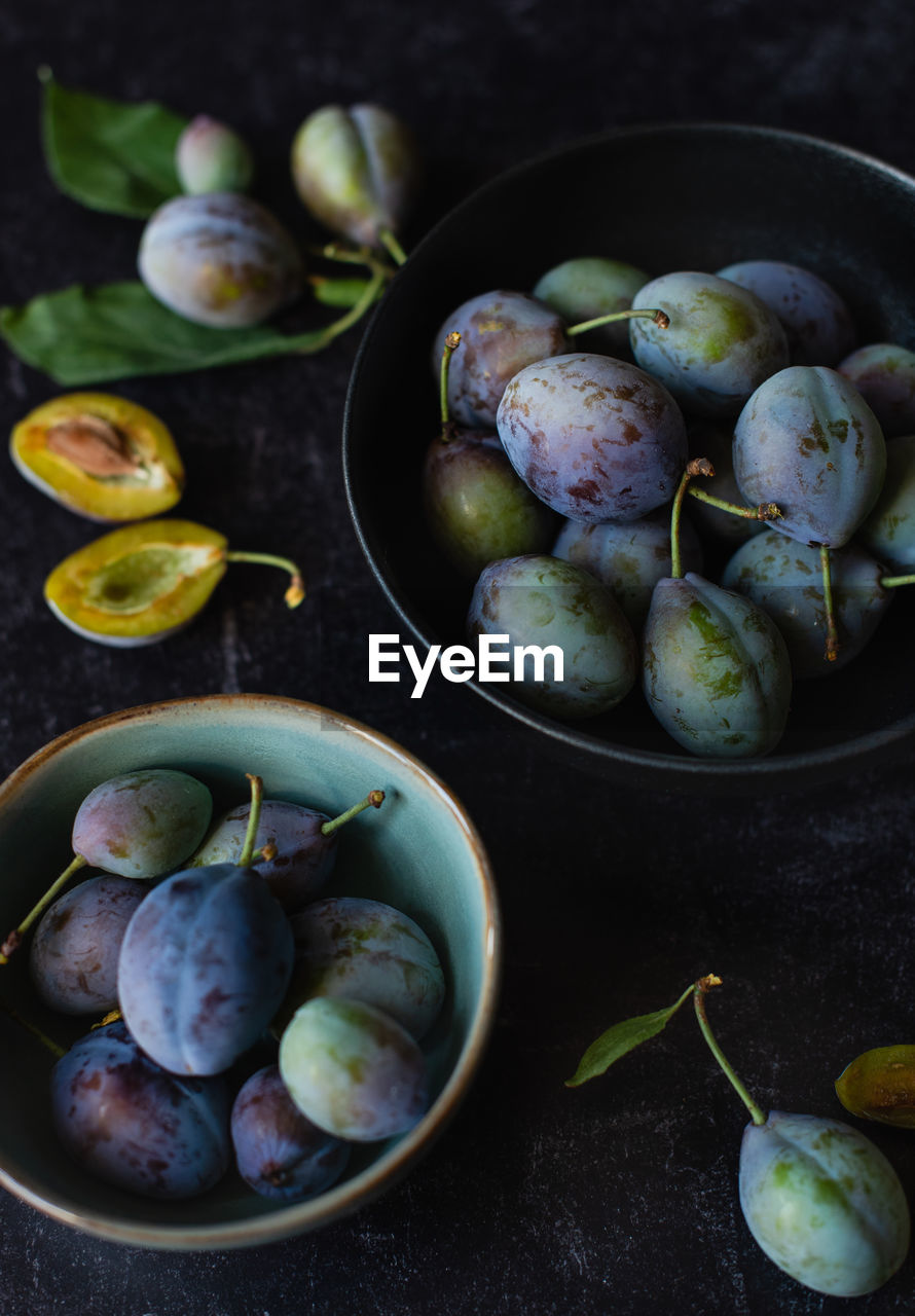 Close up of bowls of fresh plums against a black background.