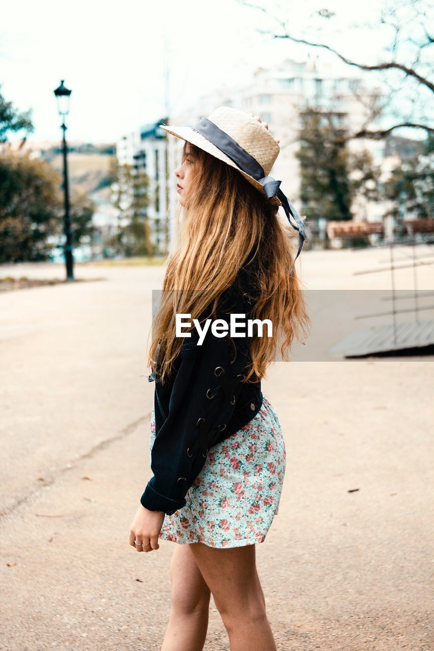 Side view of young woman wearing hat standing on road against sky