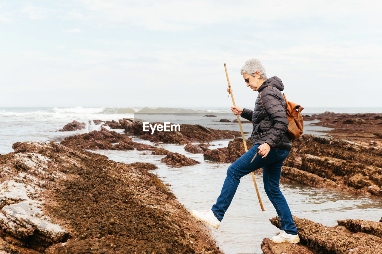 Side view of elderly female backpacker with trekking pole strolling on boulders against stormy ocean under cloudy sky