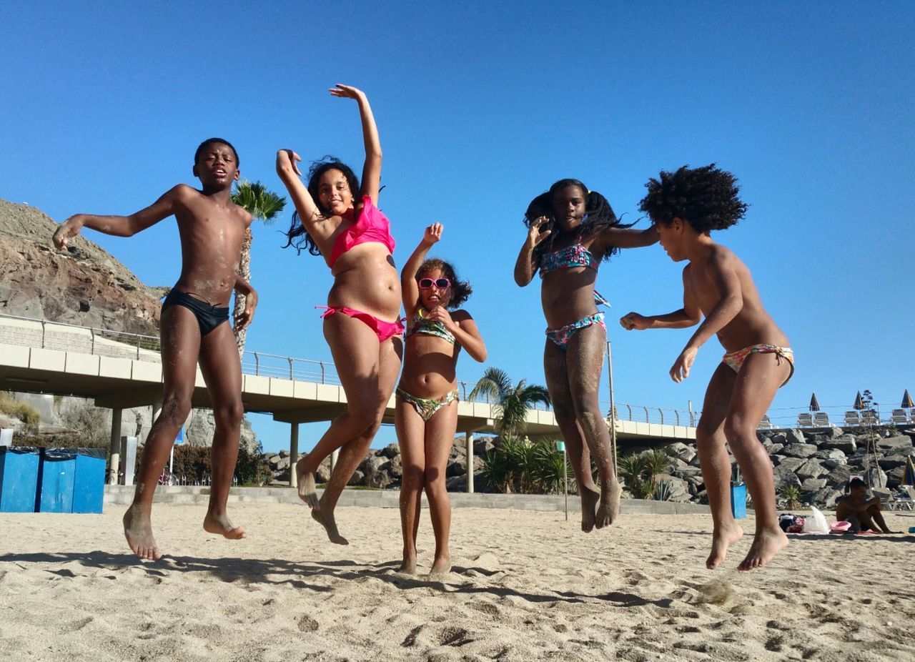 Full length of siblings jumping at sandy beach against clear blue sky