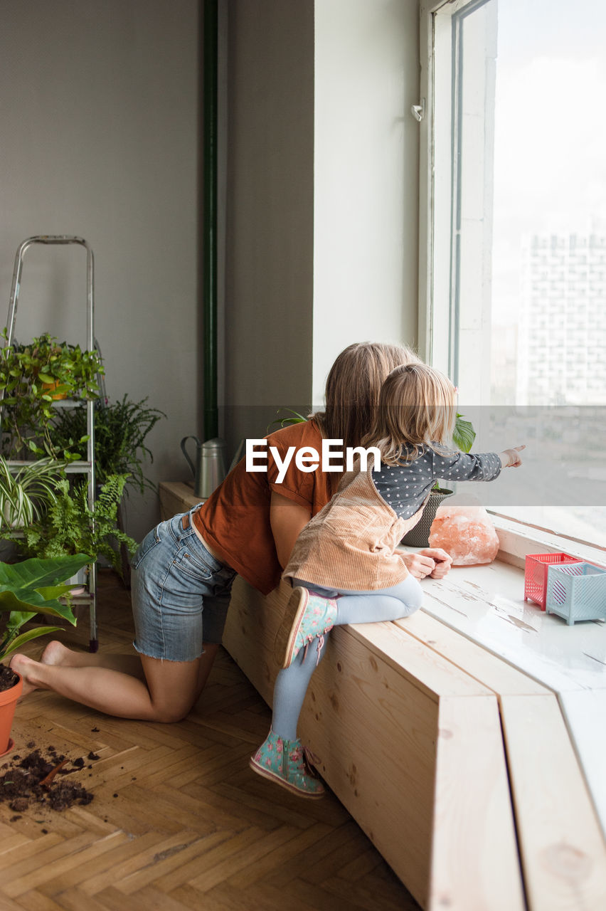 Mother and daughter look outside kneeling at window. kid points.