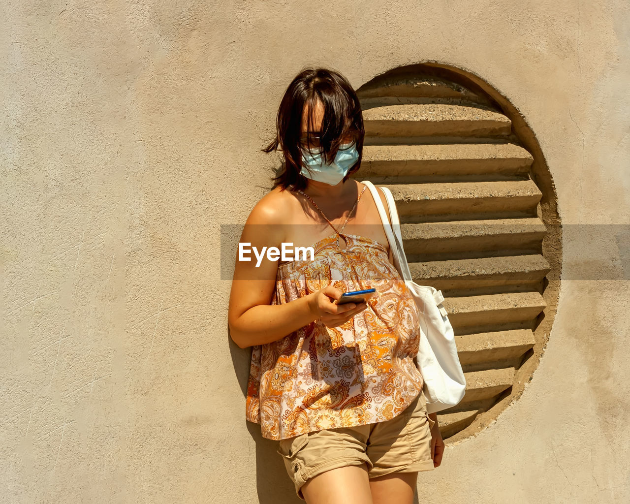 Woman using phone on a bus stop wearing surgical protective mask due to the coronavirus normative