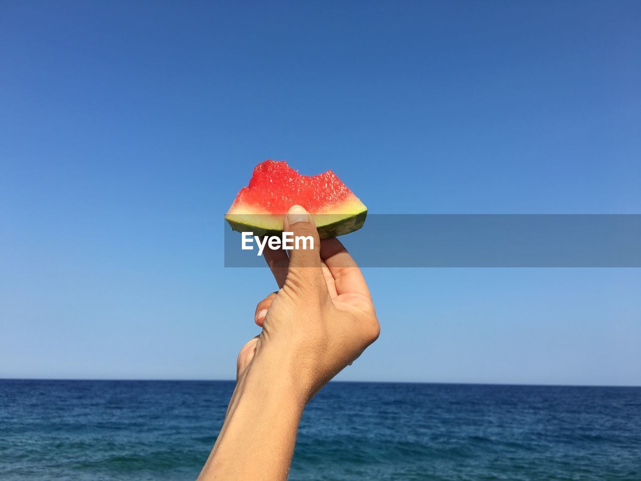 Cropped hand holding watermelon slice by sea against clear blue sky