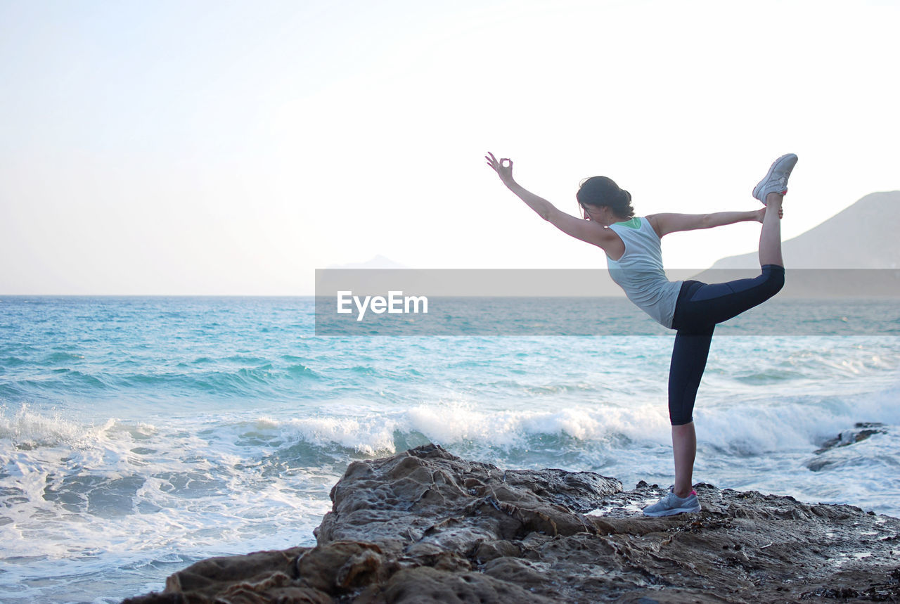 Rear view of woman doing yoga at rocks beach against clear sky 