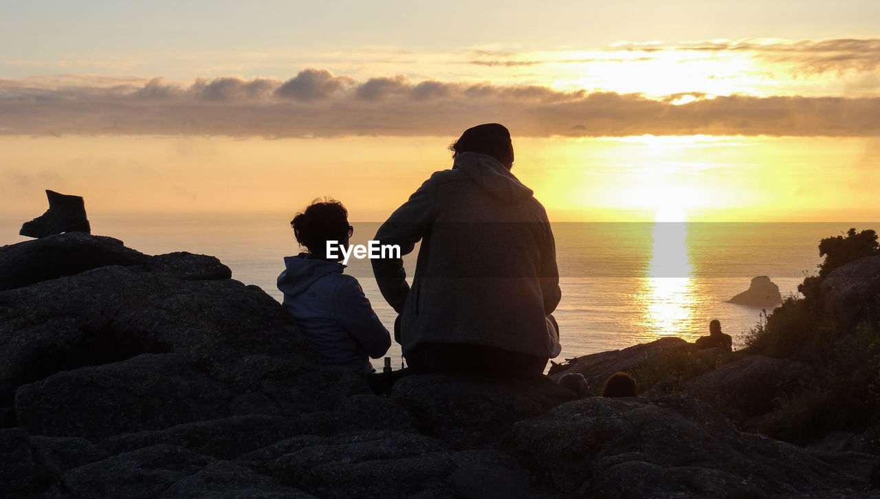 People sitting on rock by sea against sky during sunset