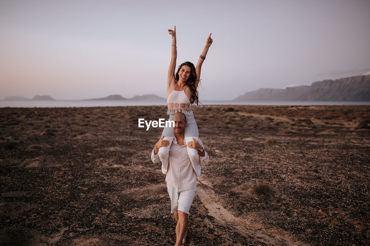 Cheerful woman with raised arms sitting on shoulders of smiling man walking along savanna in evening