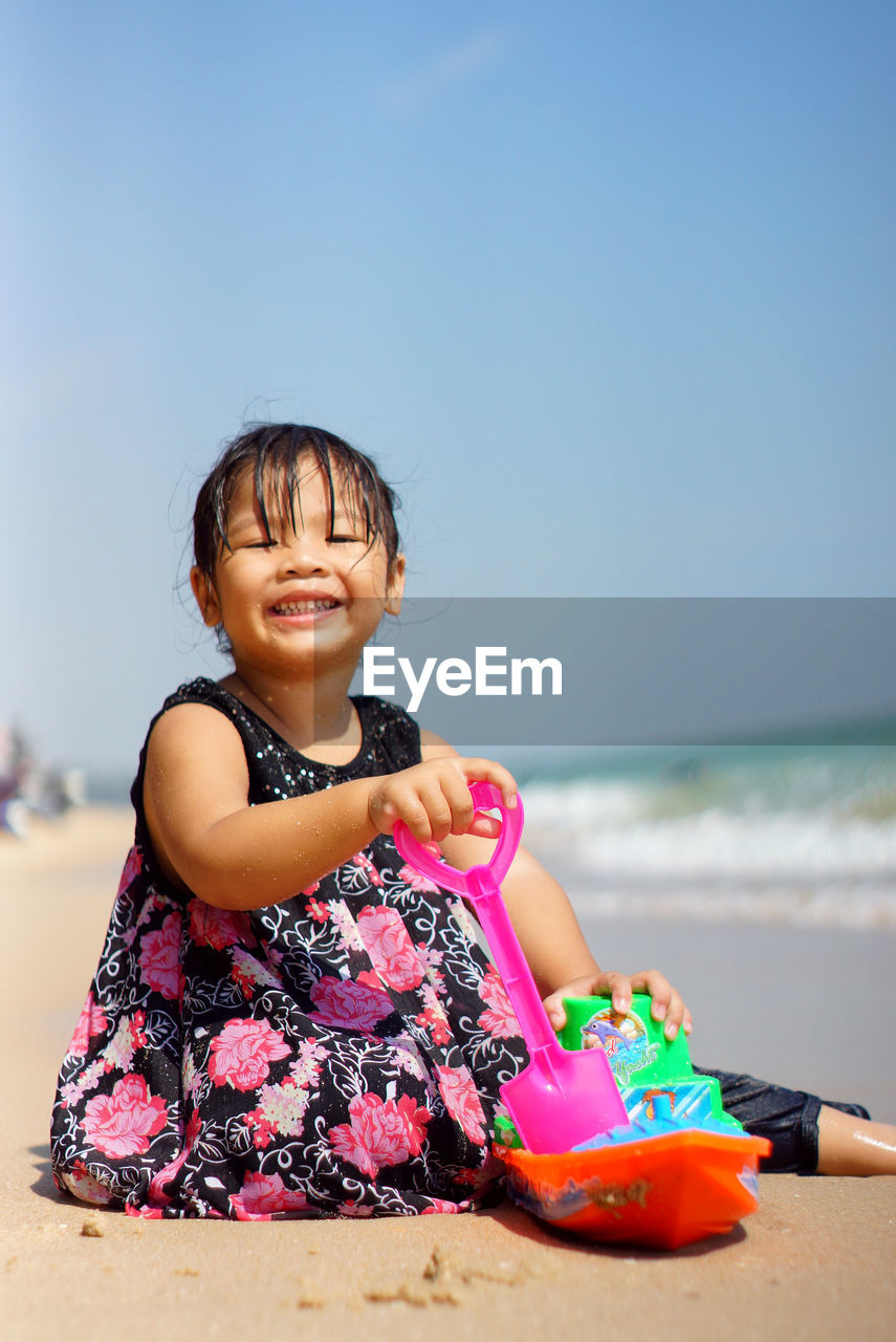 Cute smiling girl sitting with toys at beach against sky
