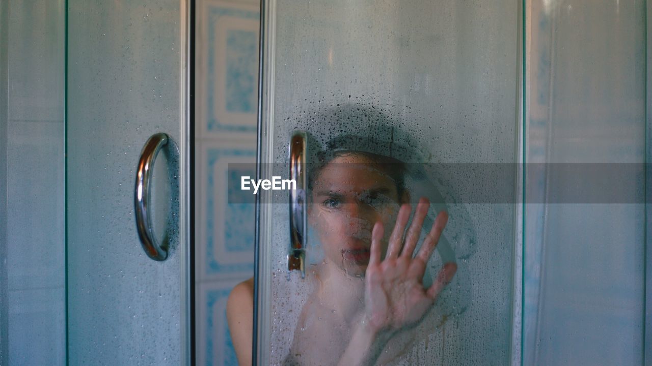 Portrait of shirtless young man looking through condensed window