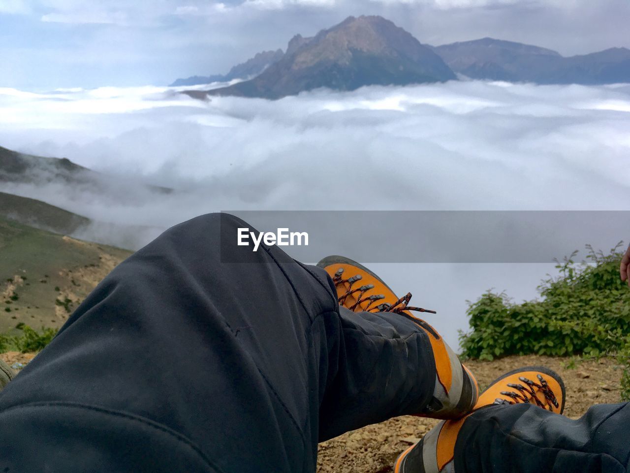 Low section of man sitting on mountain against cloudy sky