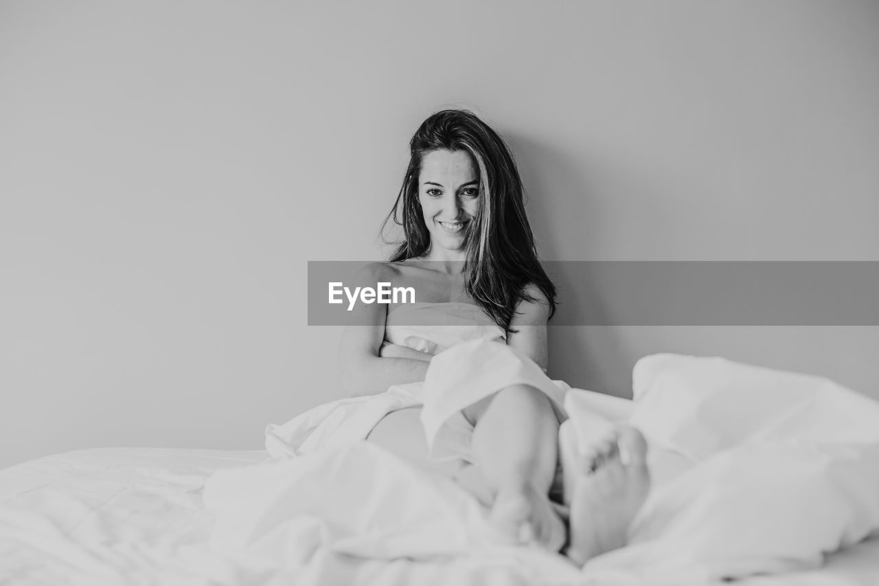 Portrait of smiling woman with blanket sitting on bed against wall