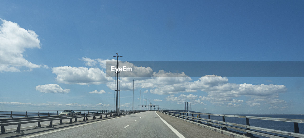 sky, transportation, road, horizon, cloud, the way forward, nature, highway, sea, water, day, diminishing perspective, railing, no people, travel, street, vanishing point, blue, symbol, beauty in nature, scenics - nature, coast, freeway, architecture, road marking, outdoors, marking, bridge, travel destinations, city, street light, sign, environment, mode of transportation, built structure, horizon over water, infrastructure, tranquility, land, copy space, tranquil scene, landscape, car, sunlight, walkway, tourism, motor vehicle, urban skyline