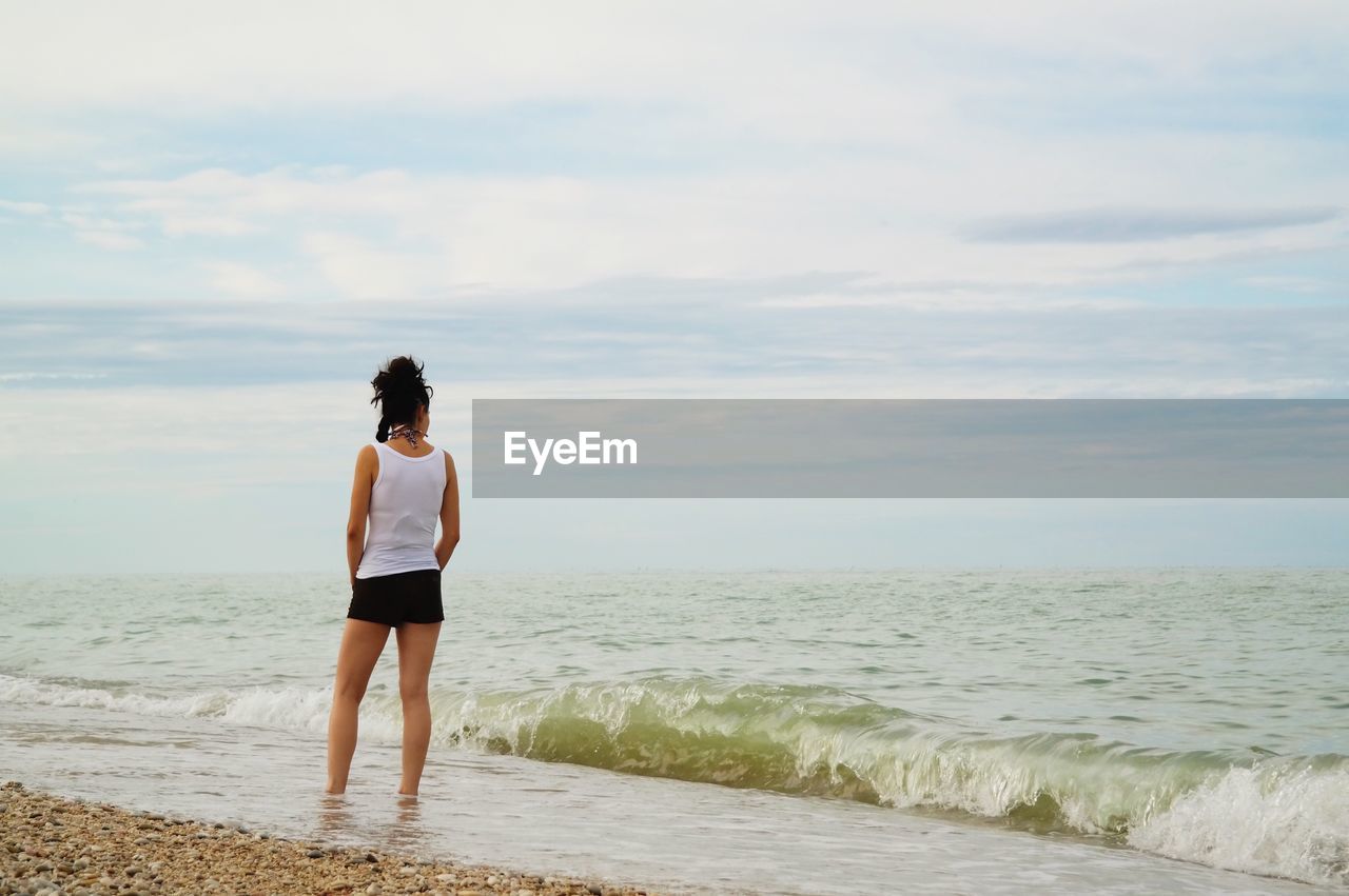 Rear view of woman standing shore at beach against cloudy sky