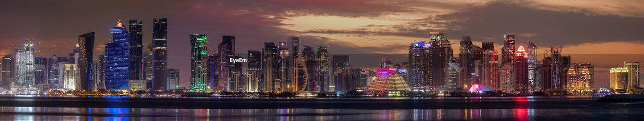 Illuminated modern buildings by river against sky at night