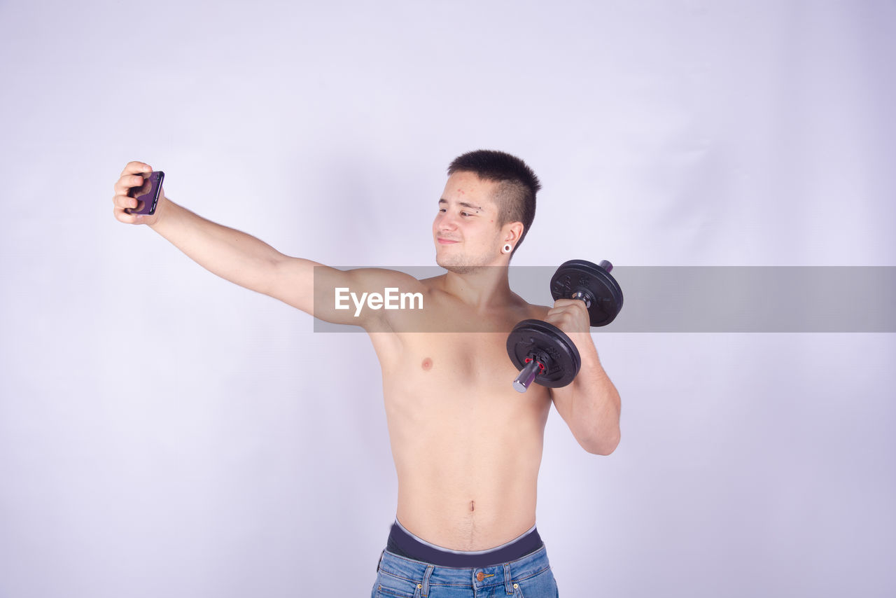 Shirtless man lifting dumbbell taking selfie with smart phone against purple background