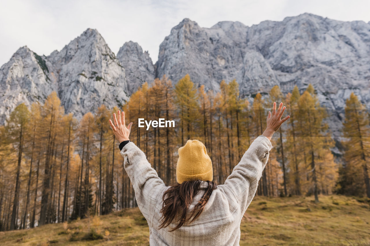 Rear view of young woman with raised arms standing under beautiful mountains in autumn