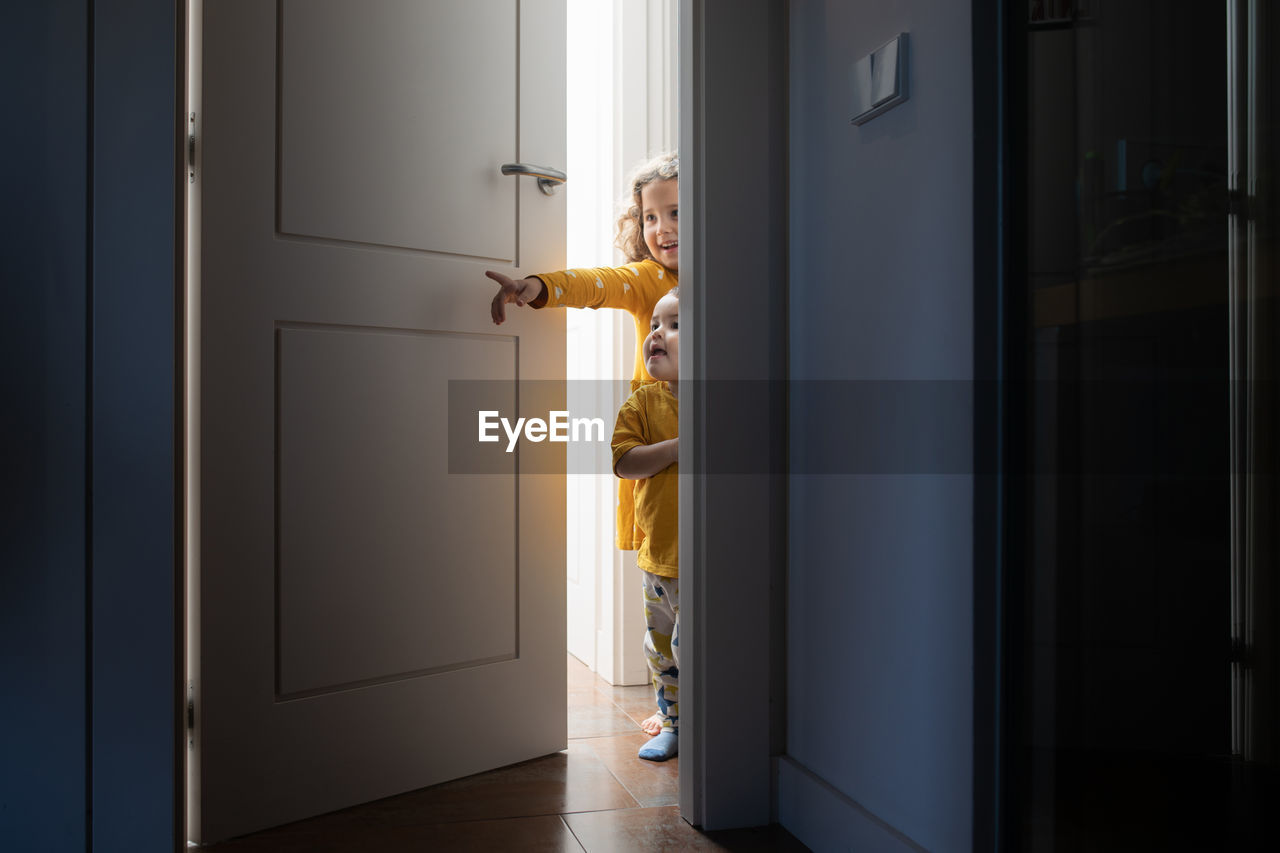 Charming brother and sister peeping out from door in modern apartment while having fun and looking away