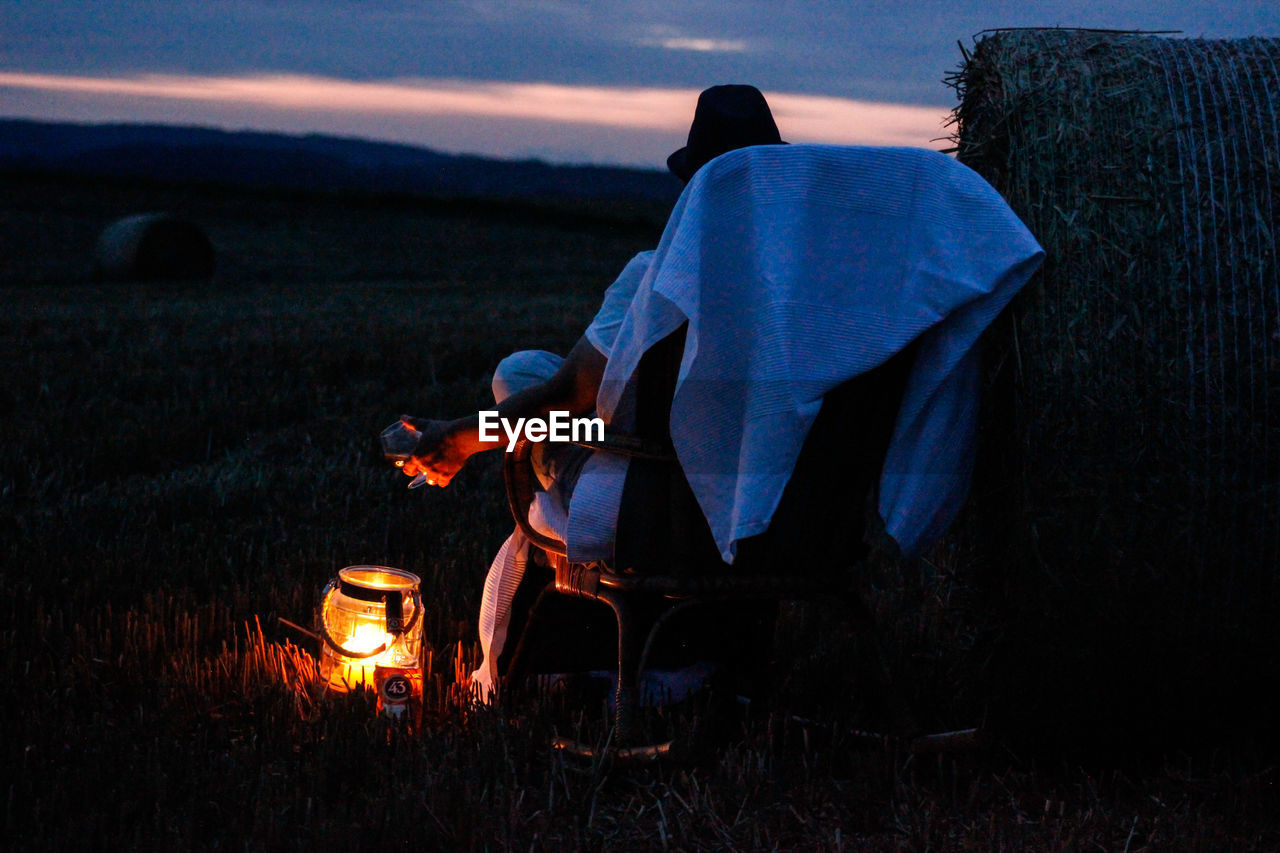 Rear view of man sitting on armchair by hay bale and lamp on farm