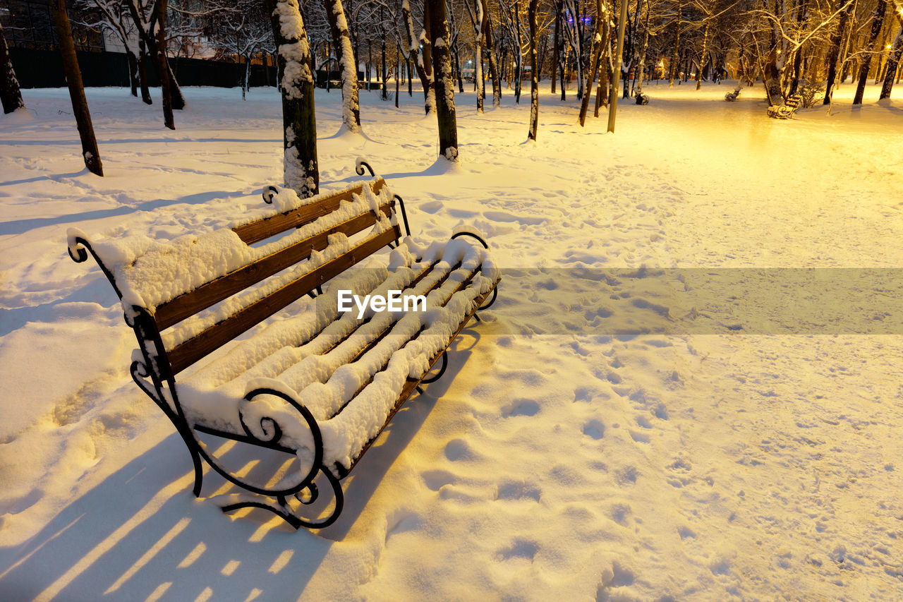 Empty brown wooden bench with metal decorative railing in the snow on winter evening in a city park.
