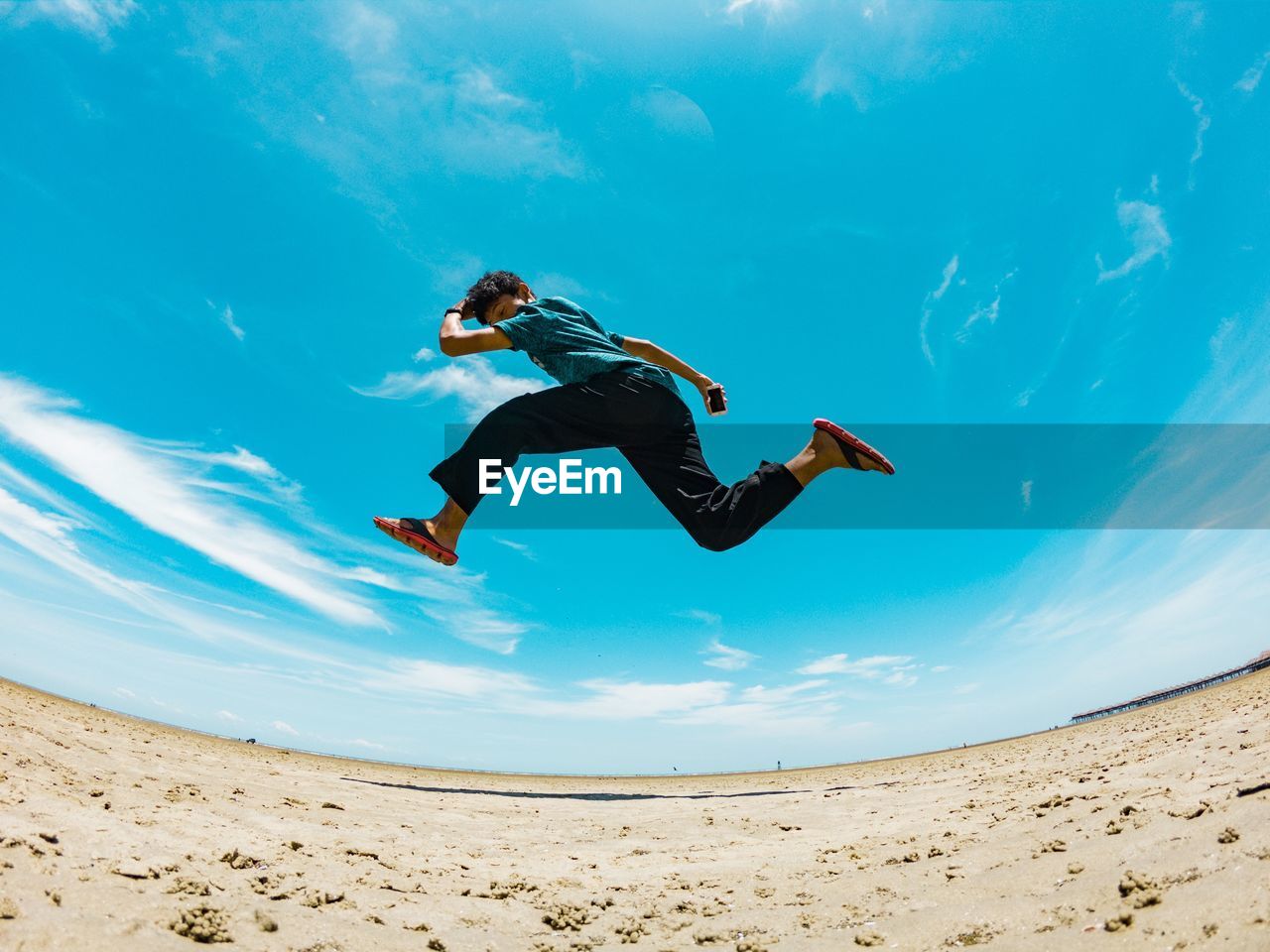 Low angle fish-eye lens shot of man jumping at beach against sky during sunny day