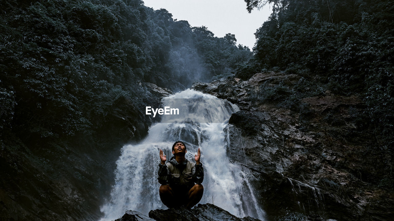 Young man meditating while sitting cross legged on rock against waterfall at forest