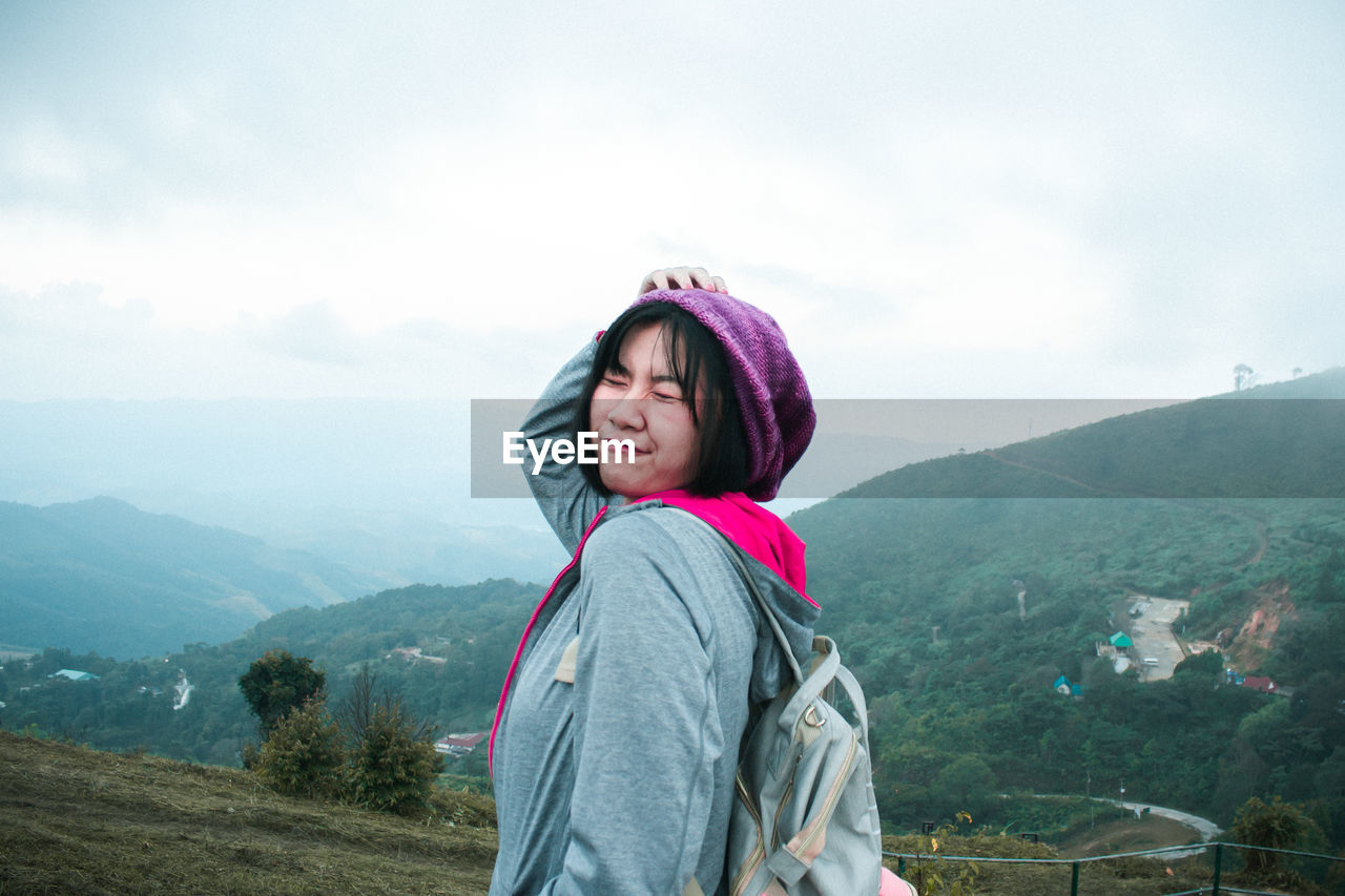 Backpacker making face while standing against mountain range against sky
