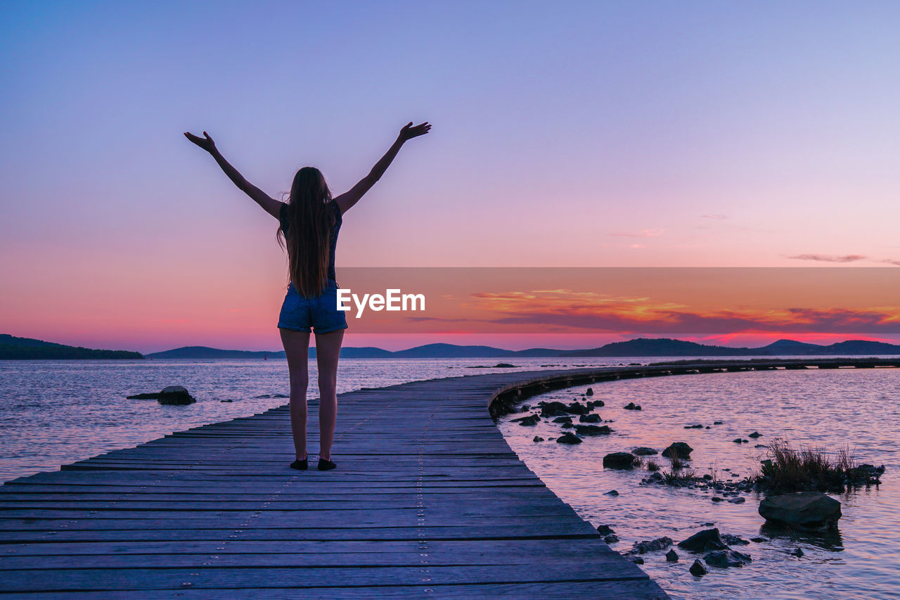 Rear view of woman standing at sea shore against sky during sunset.