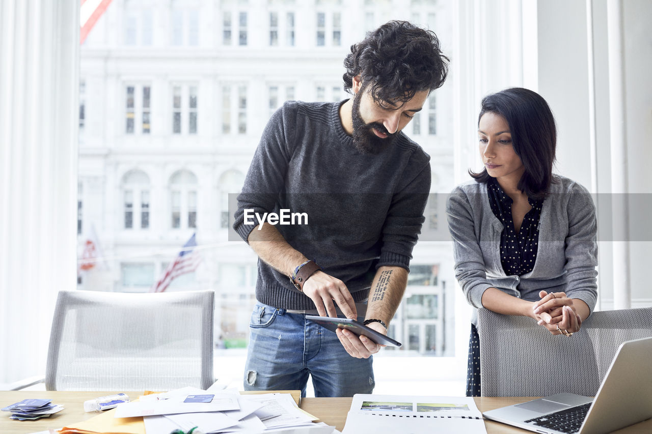 Businesswoman showing tablet computer to female colleague while standing against window in creative office