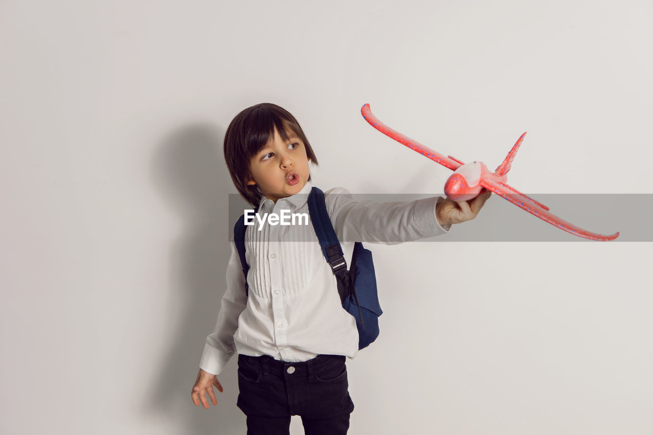 Boy child in a white shirt and jeans flies on a red airplane toy at the white wall of the house