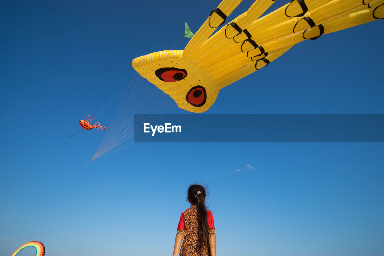 Low angle view of girl looking at kites flying in clear blue sky during festival