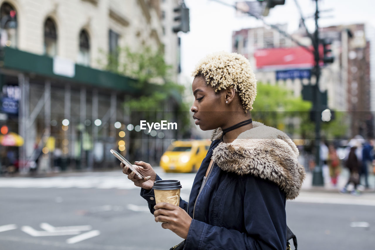 Usa, new york city, fashionable young woman with coffee to go looking at cell phone on the street