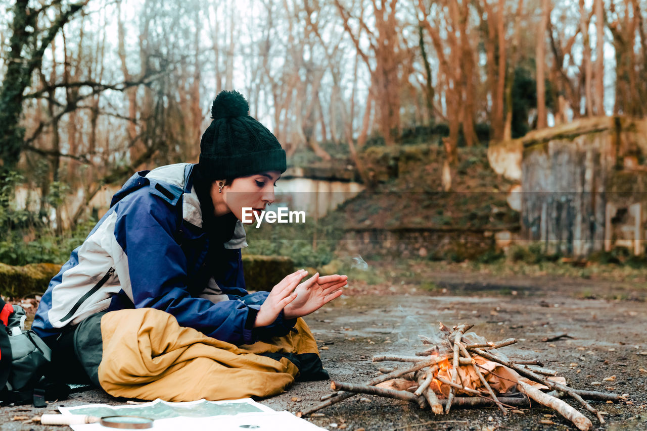 Side view of young female camper in warm clothes warming hands at bonfire while sitting on ground in countryside in autumn day