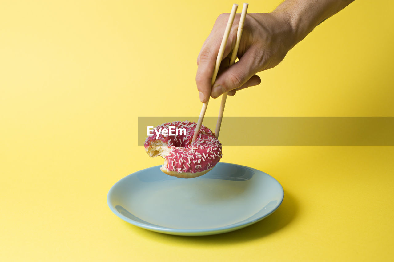 Cropped hand of person holding donuts with chopstick over yellow background