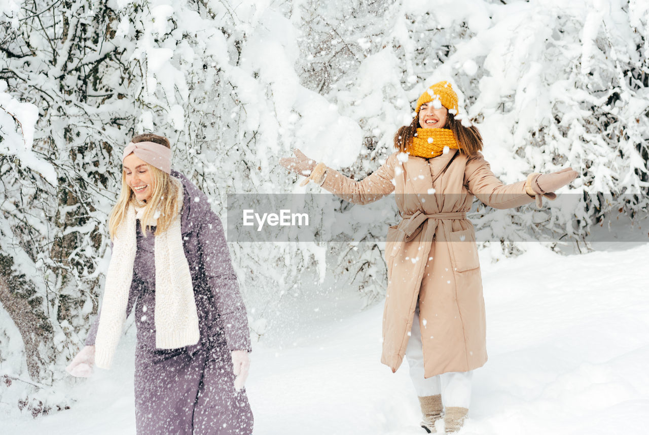 Two adult girlfriends are playing snowballs in a snowy forest and laughing.