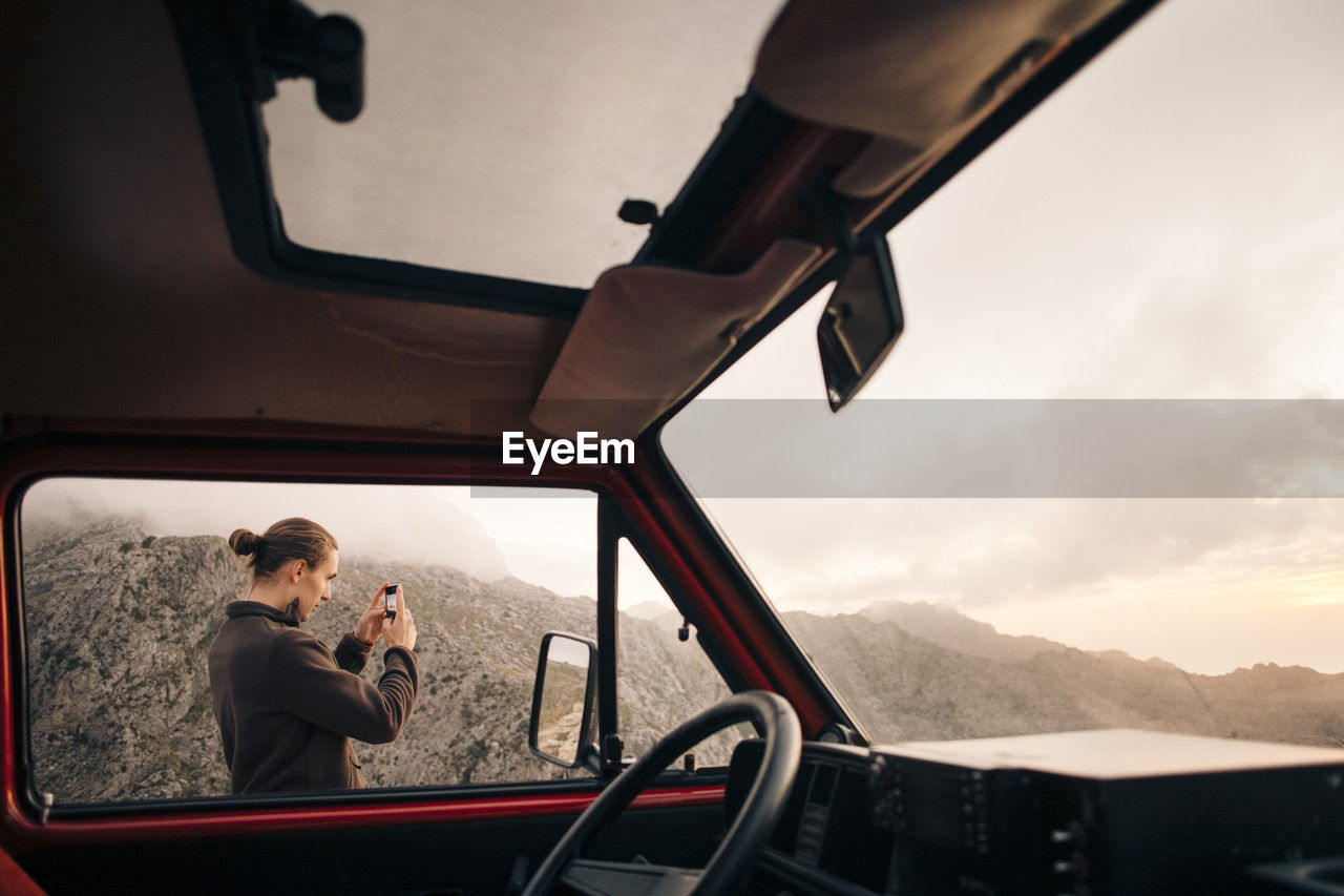 Young man photographing mountains through smart phone seen from van window on vacation