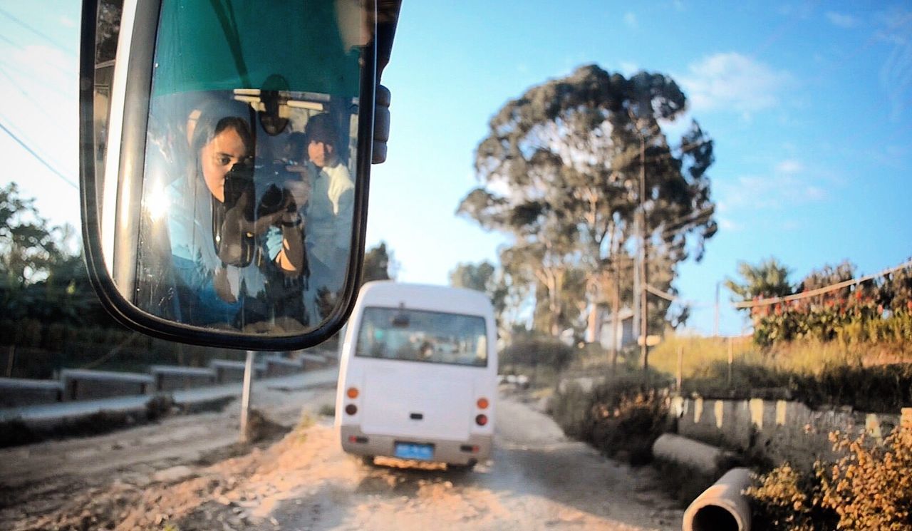 Reflection of woman with camera on side-view mirror in bus on road