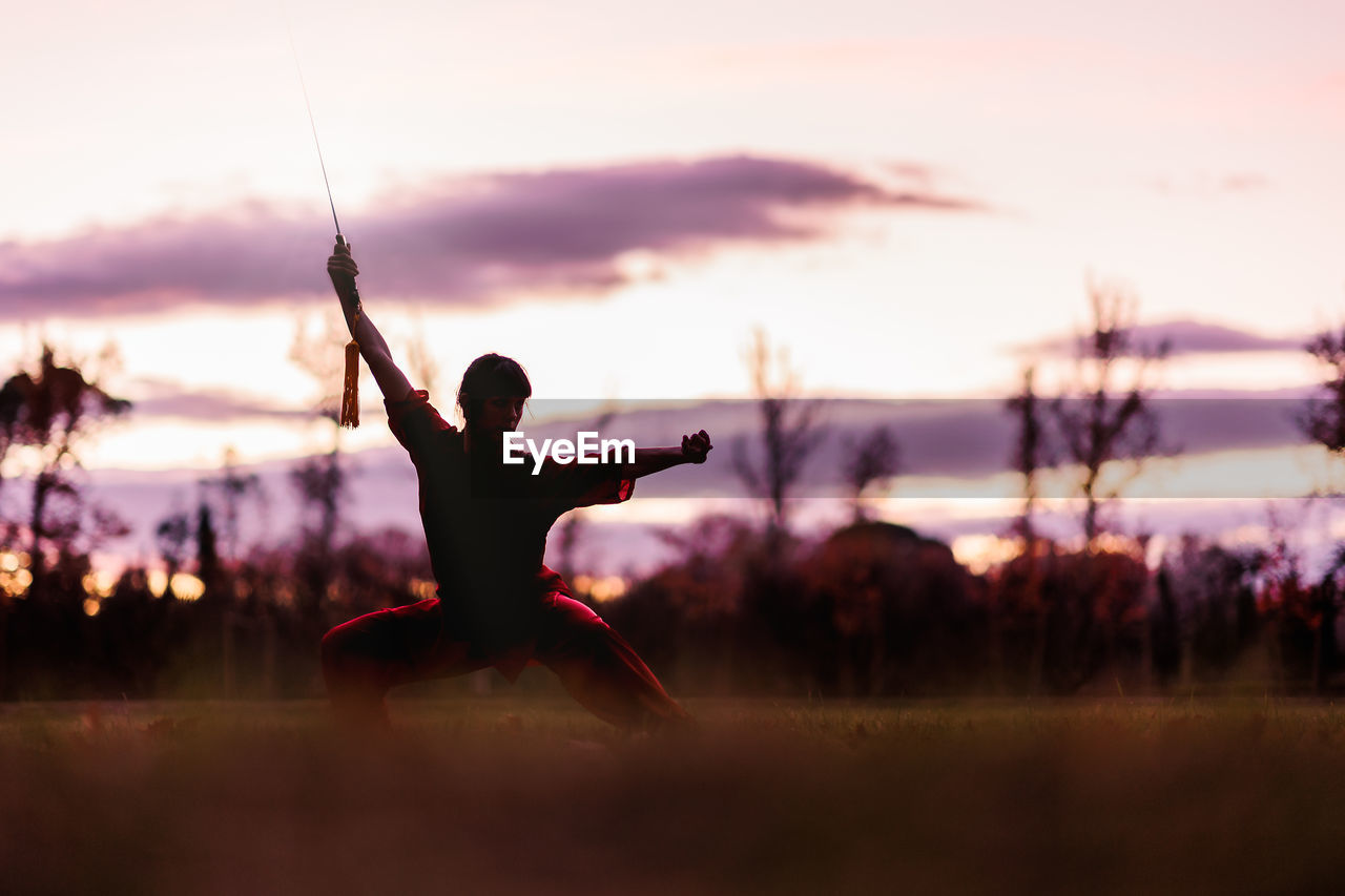 side view of woman with arms outstretched standing on field against sky during sunset