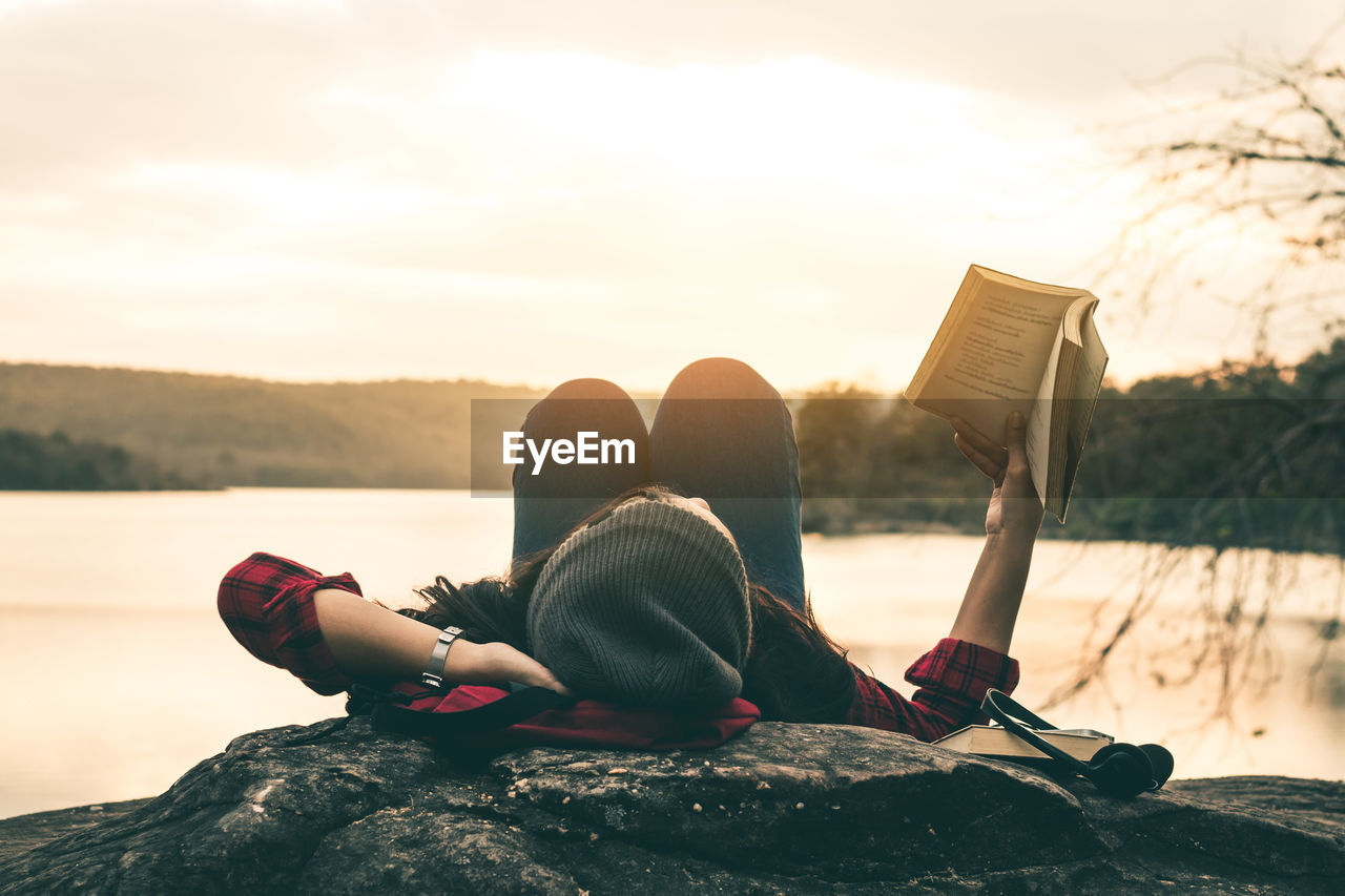 Woman reading book while lying on lakeshore during sunset