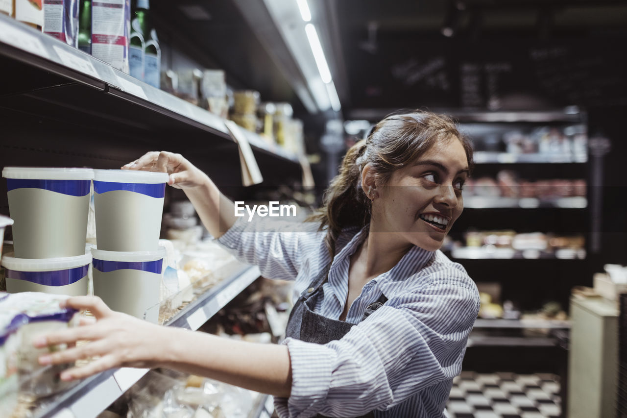 Young female owner looking over shoulder while arranging product on rack at delicatessen shop