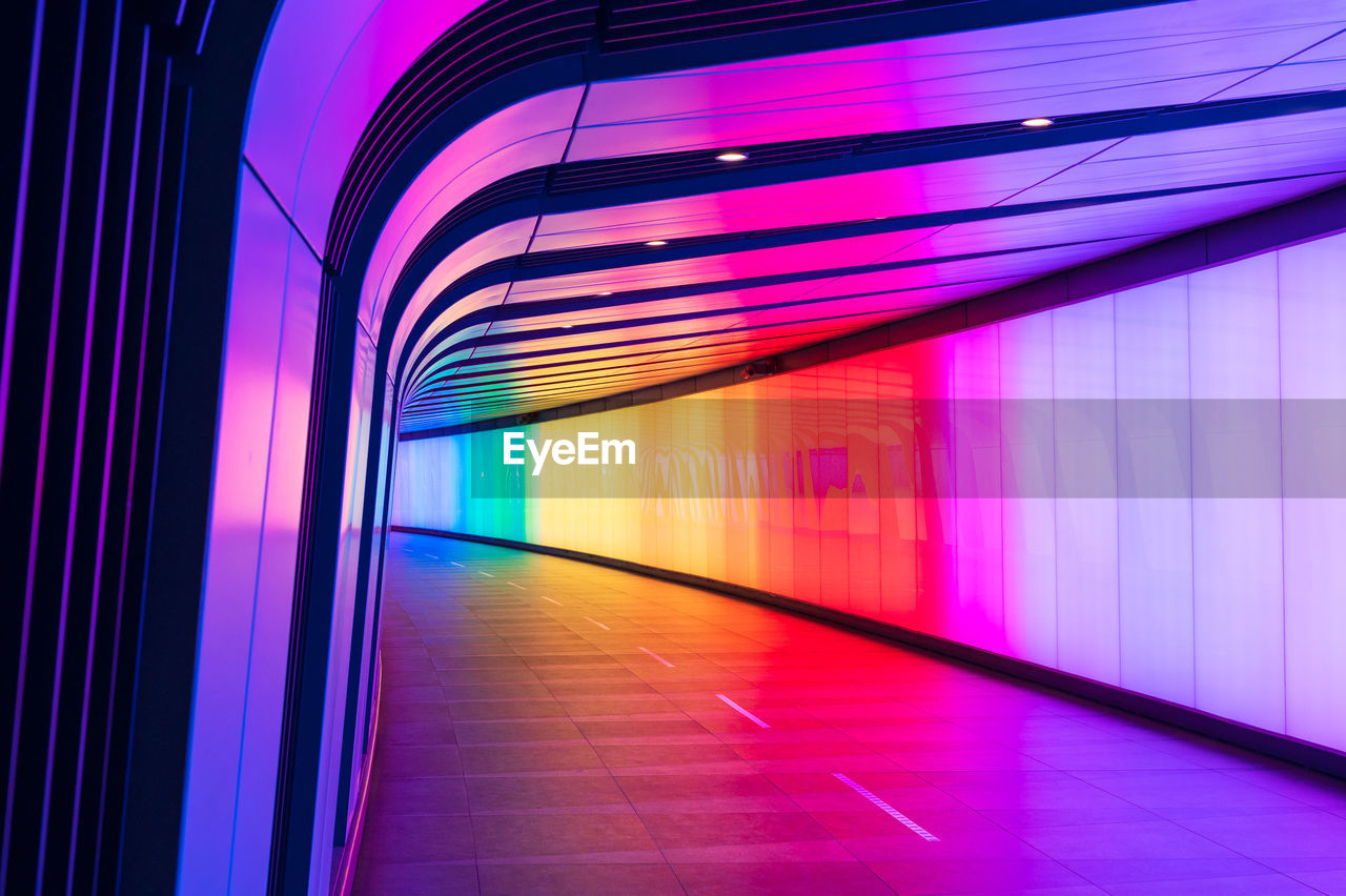 architecture, light, illuminated, multi colored, line, lighting equipment, built structure, indoors, no people, the way forward, transportation, corridor, futuristic, city, tunnel, arcade, night, purple, neon, pink, light - natural phenomenon, abstract, diminishing perspective, motion, flooring, building, speed, vibrant color, blurred motion, footpath, long exposure, wall - building feature, pattern, blue