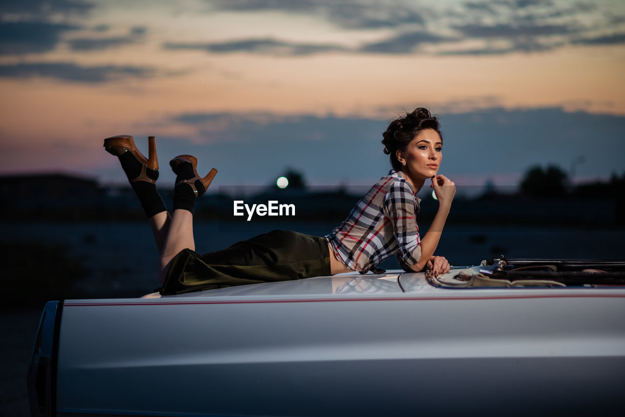 Woman sitting on car against sky during sunset