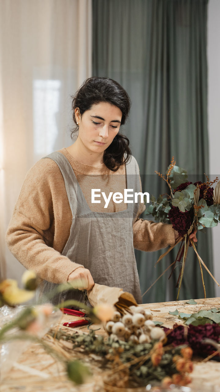 Diligent adult woman in casual clothes and gray apron making beautiful flower composition with dry green and burgundy plants in combination with brown stems while standing at table at home