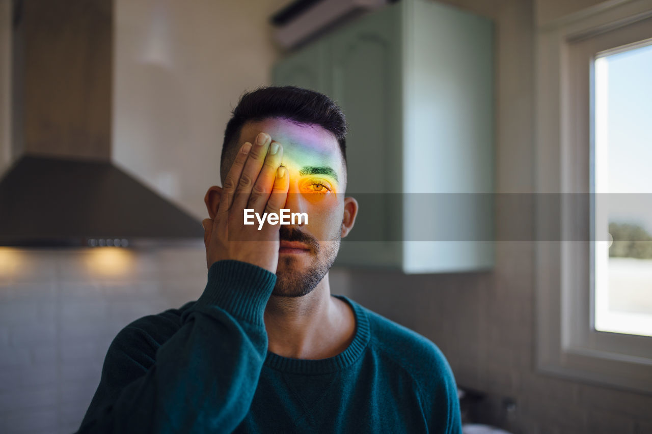 Rainbow light hitting face of young man covering one eye with hand