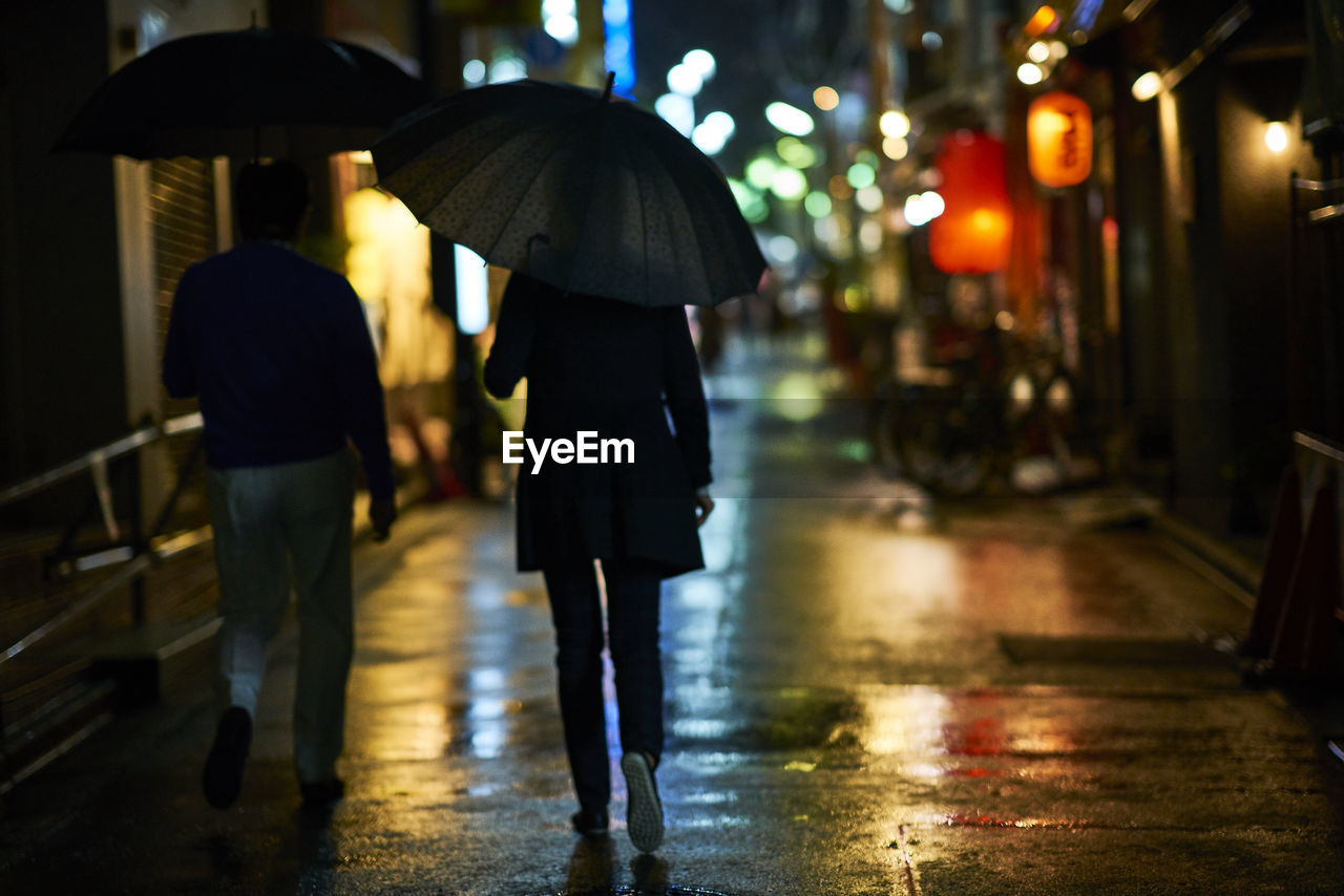 Rear view of people carrying umbrella while walking on wet street at night
