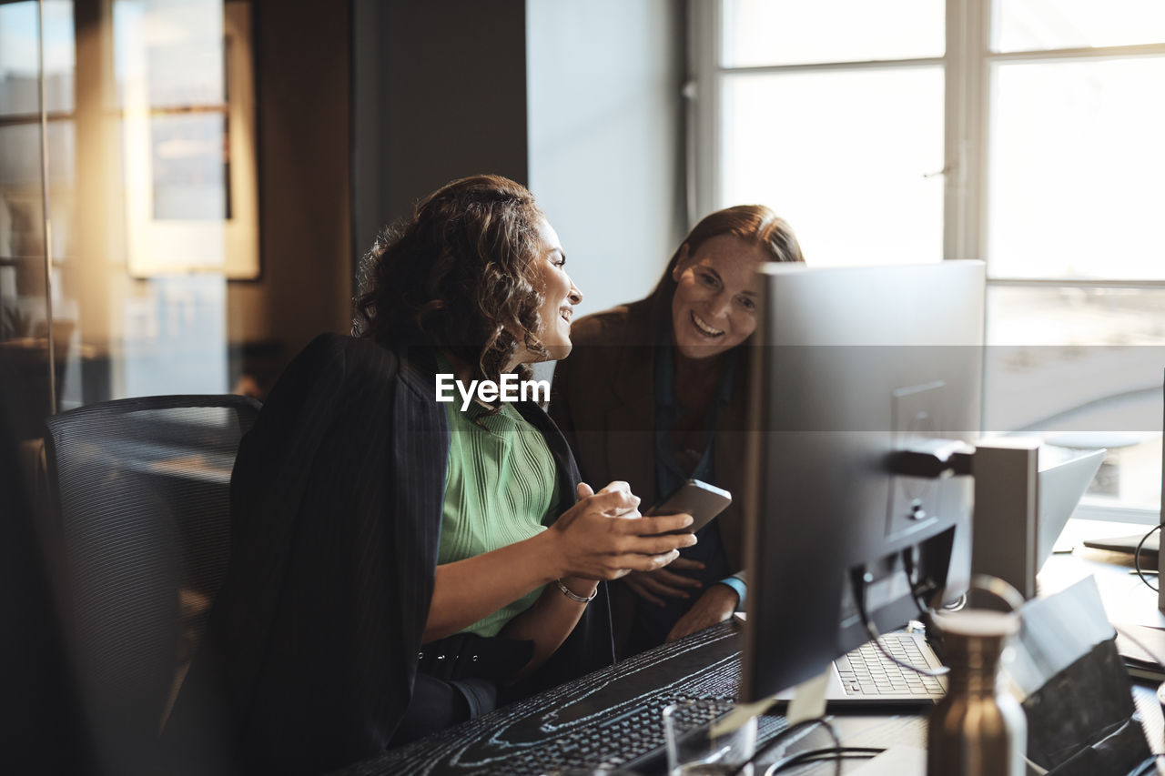 Happy businesswoman sharing smart phone with female entrepreneur sitting at desk in office