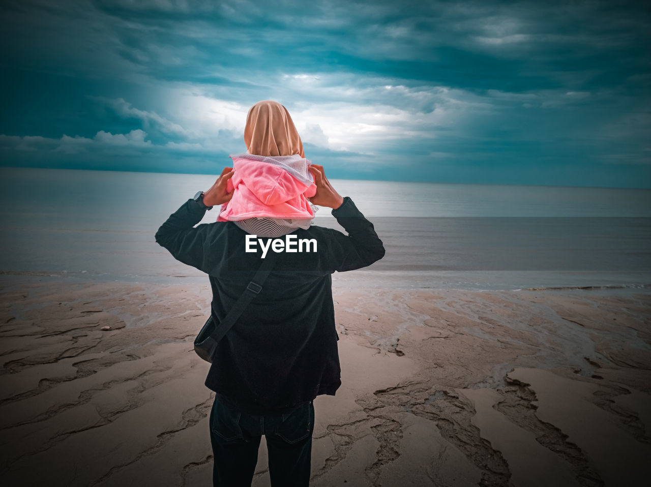 Rear view of father is holding his child standing on beach against sky